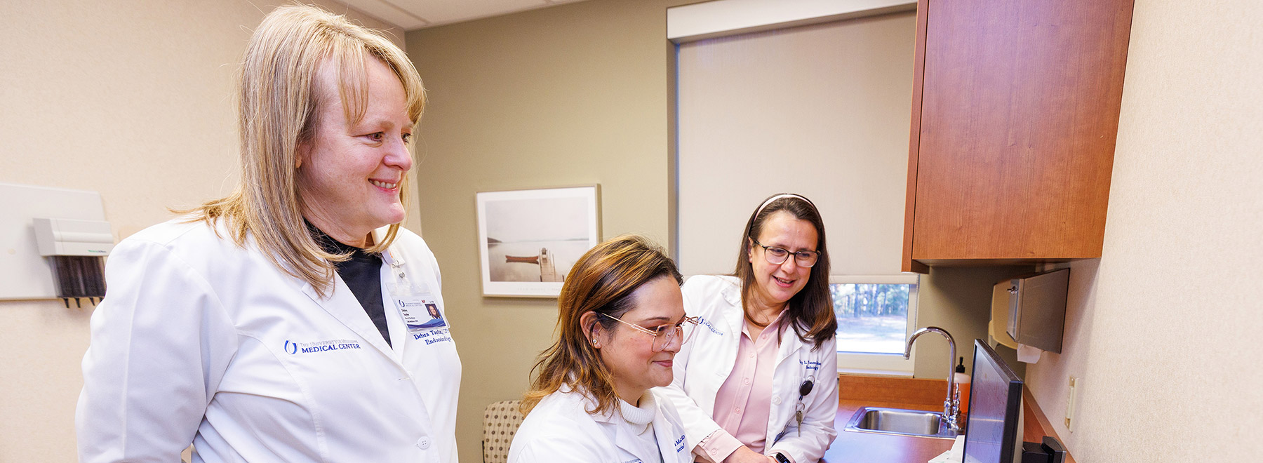 Three physicians review data on a computer monitor. One is standing, one is sitting, and one is leaning on a counter nearby.
